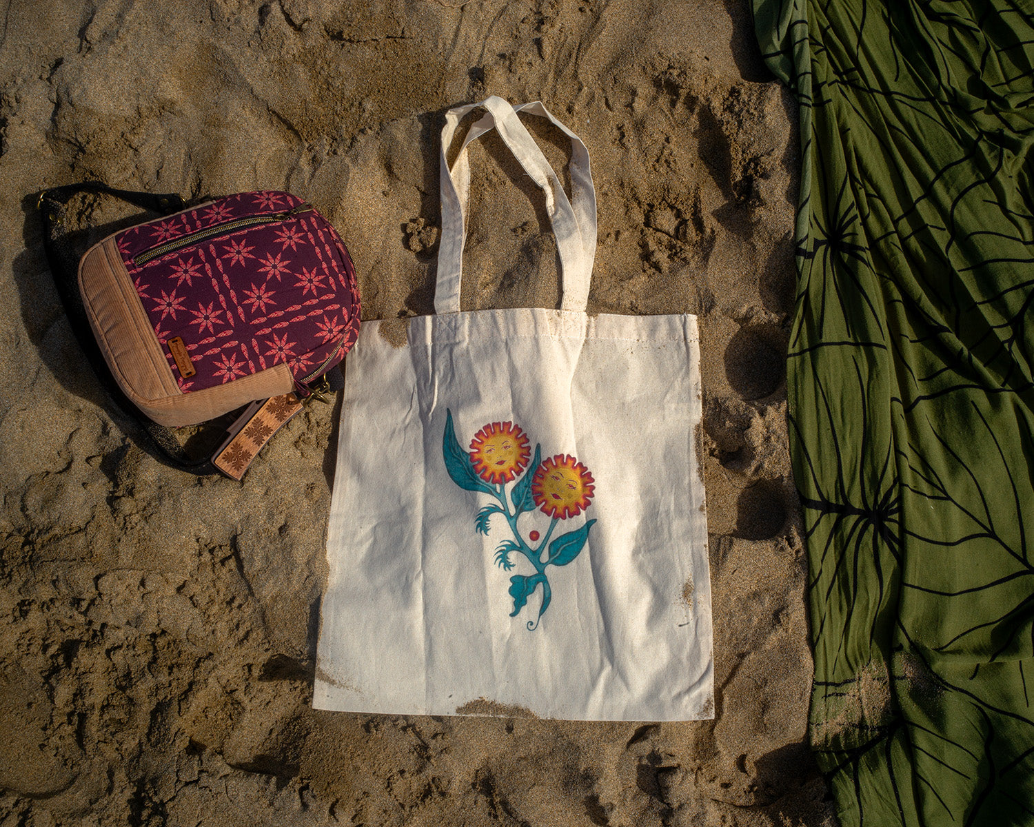 White canvas tote bag featuring an original floral painting titled “Sisters of the Stem,” showing two flowers with feminine faces growing from a single stem, photographed on sand at the beach.