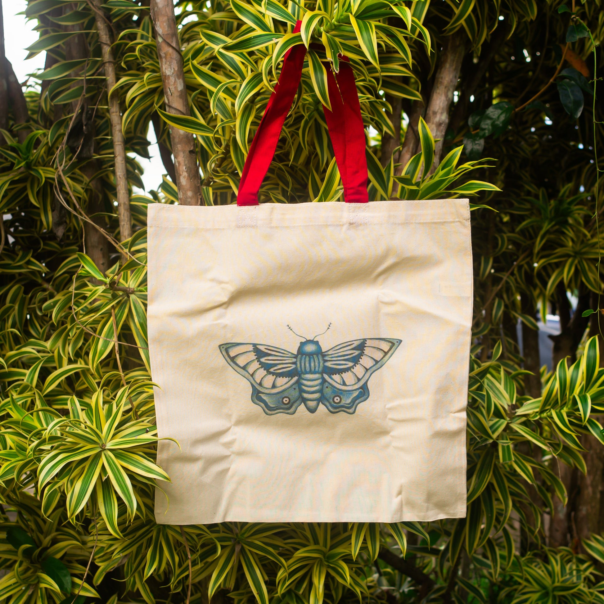Cream canvas tote bag with red handles featuring a blue butterfly illustration, hanging from a leafy tropical tree with green and yellow foliage in natural outdoor light.