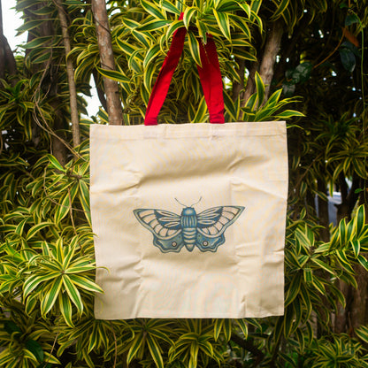 Cream canvas tote bag with red handles featuring a blue butterfly illustration, hanging from a leafy tropical tree with green and yellow foliage in natural outdoor light.