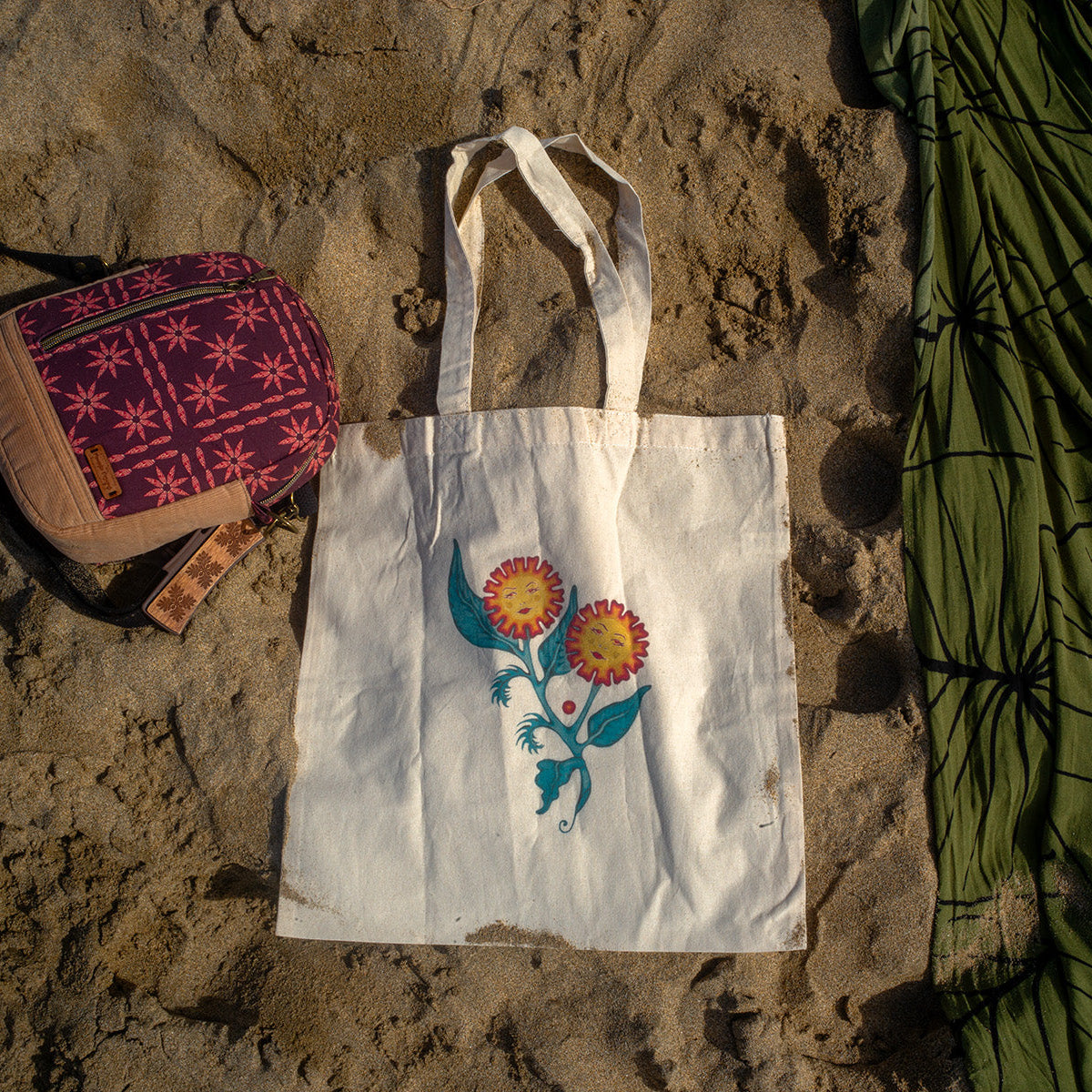 White canvas tote bag featuring an original floral painting titled “Sisters of the Stem,” showing two flowers with feminine faces growing from a single stem, photographed on sand at the beach.