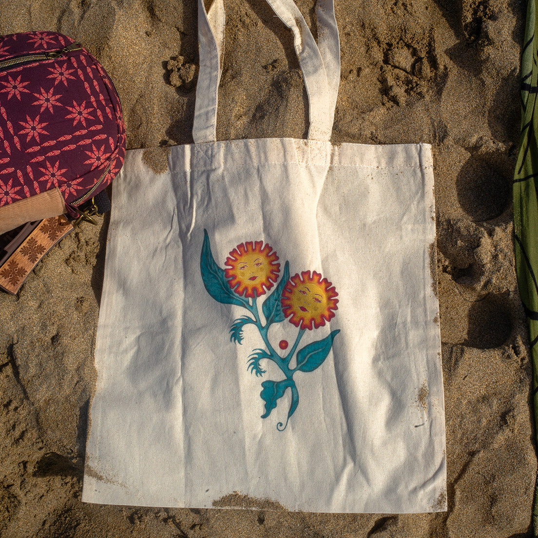 White canvas tote bag featuring an original floral painting titled “Sisters of the Stem,” showing two flowers with feminine faces growing from a single stem, photographed on sand at the beach.