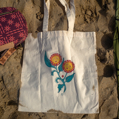 White canvas tote bag featuring an original floral painting titled “Sisters of the Stem,” showing two flowers with feminine faces growing from a single stem, photographed on sand at the beach.